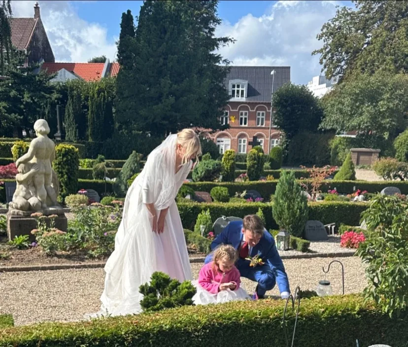 Pernille i brudekjole og Kenneth i blåt jakkesæt ved Neyas gravsted på bryllupsdagen, med en af Neyas lillesøstre ved stenen.